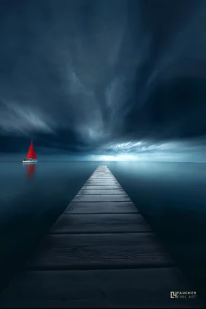 Jetée en bois menant vers l’horizon sur une mer calme sous un ciel sombre et dramatique, avec un voilier à voile rouge au loin.