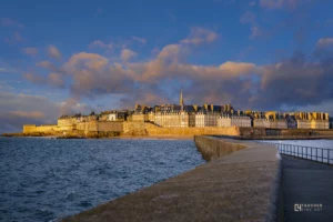 Vue de Saint-Malo intra-muros au coucher du soleil, remparts illuminés, mer agitée et ciel nuageux aux teintes dorées, photographie Fine Art.